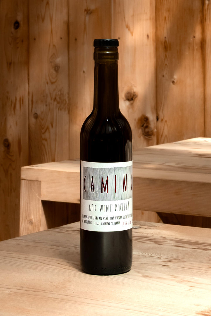 A dark glass bottle labeled Camino Red Wine Vinegar from Broc Cellars stands on a light wooden surface with a wooden wall and bench behind. Warm, natural light highlights this unique organic vinegar.