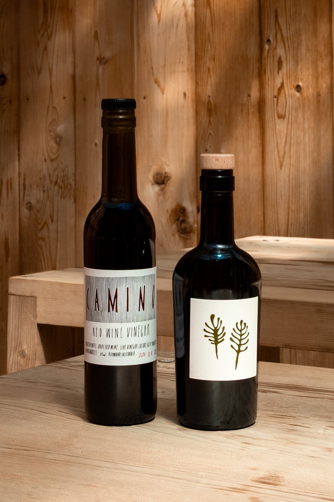 Two dark glass bottles—a Broc Cellars Camino Vinegar and Fox Hill Extra Virgin Olive Oil Duo—sit on a wooden surface with a simple plant drawing on the oil label, making this pair an essential kitchen staple.