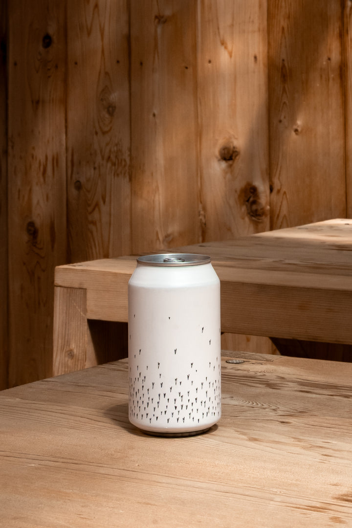 A Broc Cellars Love Sparkling Can Pack, featuring a white can with a minimal black dotted pattern, sits on a wooden table. Wooden panels and another table are visible in the naturally lit background.