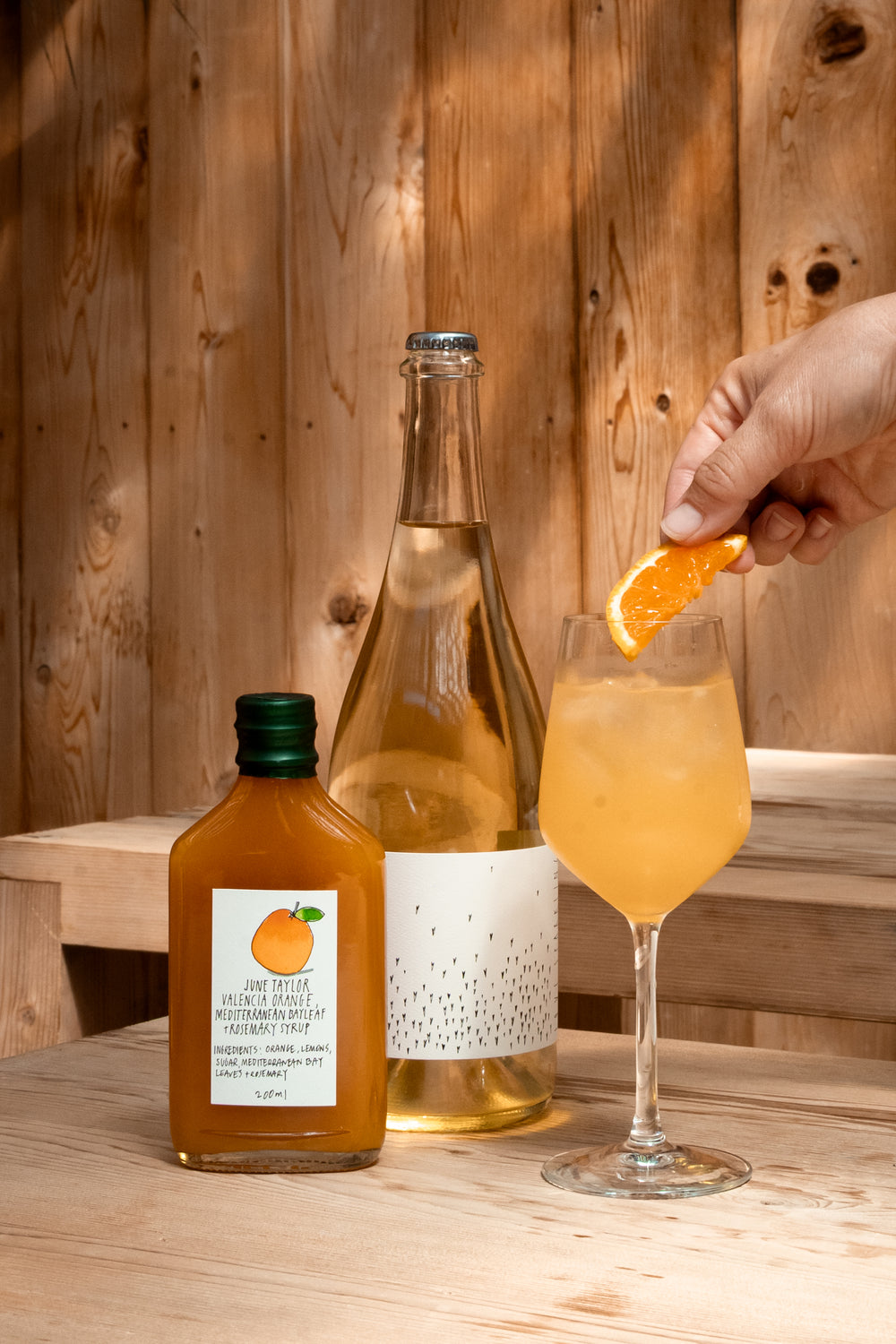 A hand places an orange slice into a glass of orange drink on a wooden table, next to a Broc Cellars Spritz Pack with June Taylor Seasonal Fruit Syrup. A wooden wall is in the background.