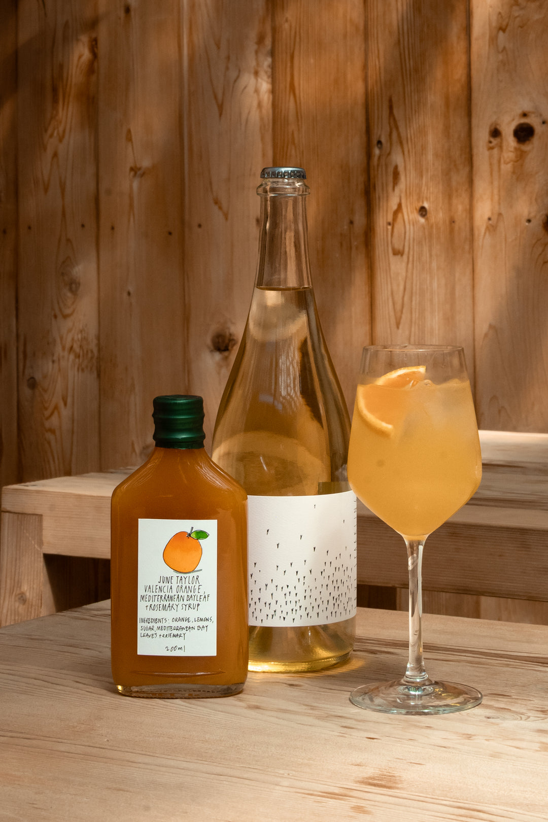A glass of orange cocktail with citrus, a clear bottle, and a small June Taylor Seasonal Fruit Syrup from the Broc Cellars Spritz Pack sit on a wooden table in front of a wooden wall.