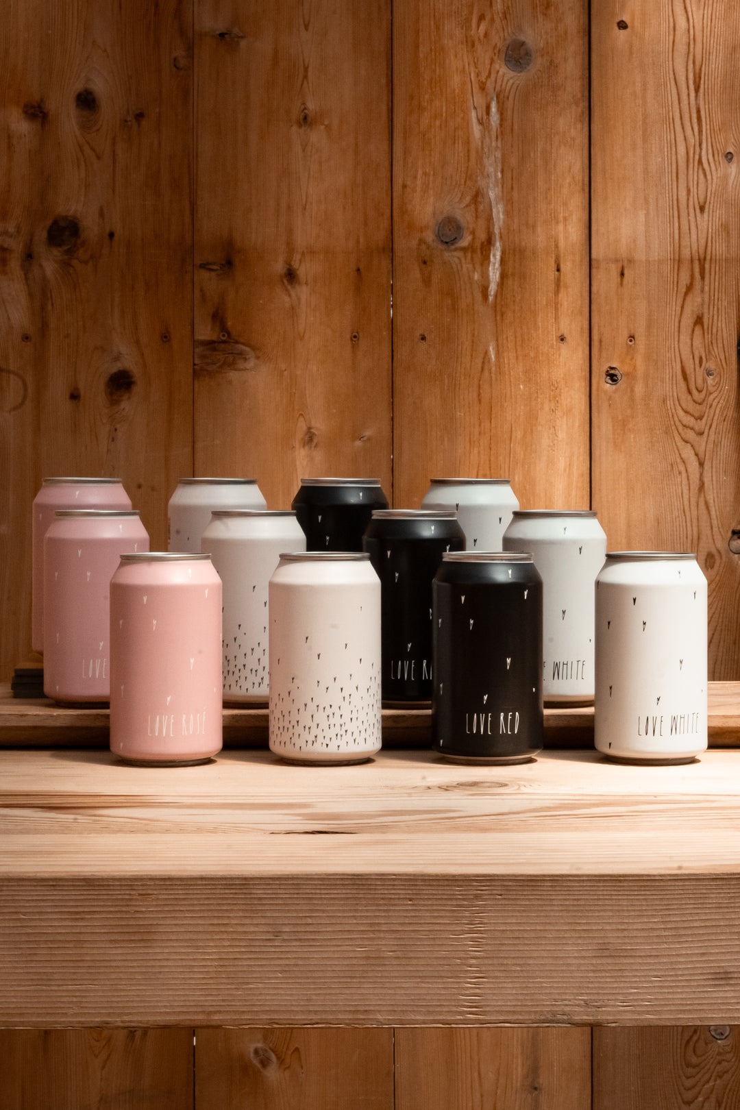 A selection of Broc Cellars’ Love Can Variety Pack in pink, white, black, and speckled cans sits on a wooden shelf. The canned wine features minimalist text and design—ideal for stylish picnic drinks.