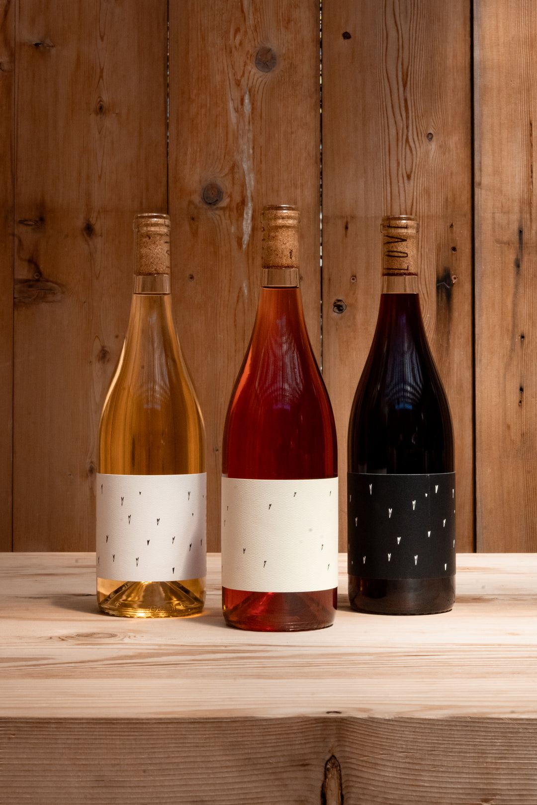 Three Broc Cellars Love Trio wine bottles—white, rosé, and red—stand on a light wooden surface with minimalist labels, making this set ideal for parties or as an elegant gift.