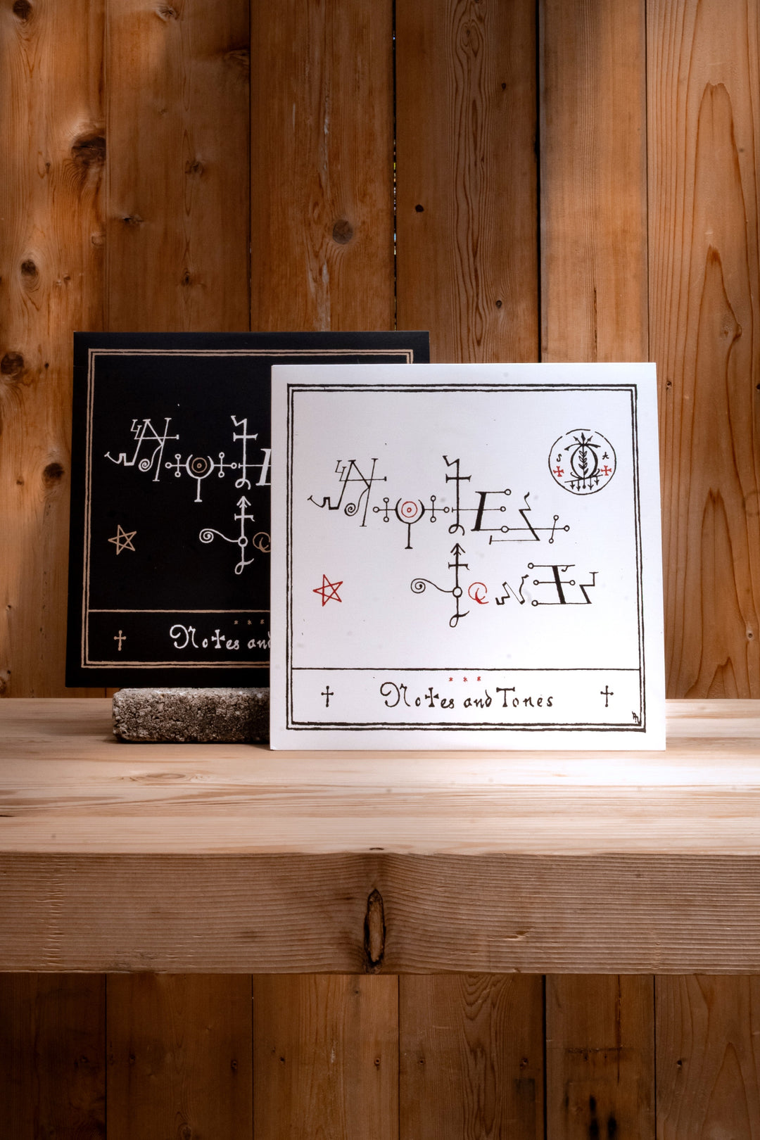 Two square cards from Broc Cellars' Notes & Tones V.1 + V.2 Album Duo rest on a wooden shelf—one white with black and red marks, the other channeling DJ Muggs’s vibe as Sun Ra samples echo from the wood-paneled wall behind.