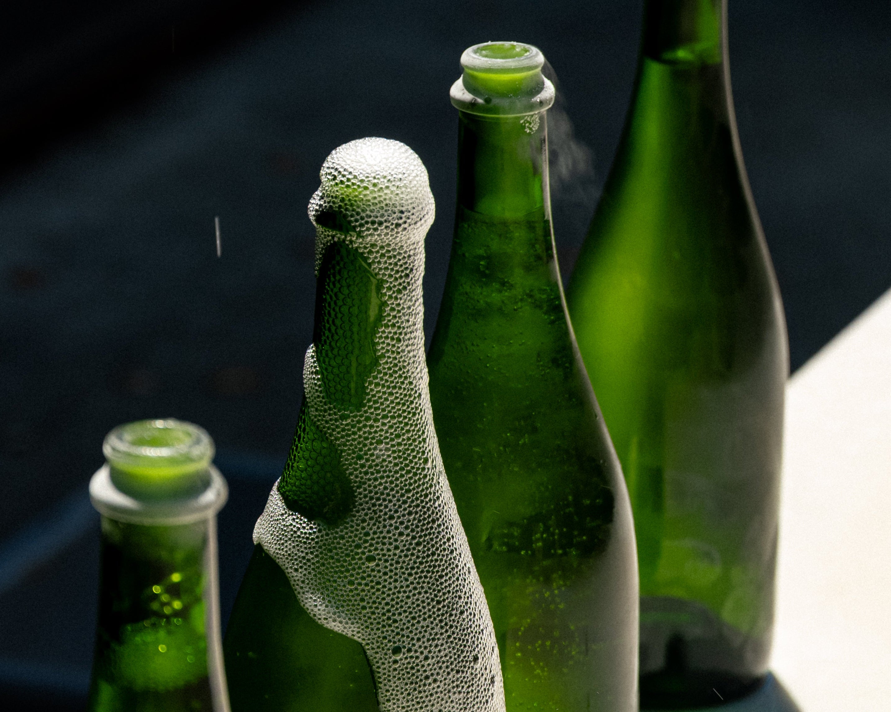 Three green glass beer bottles with condensation and foam, photographed close up on a dark surface with a bright white area in the background.