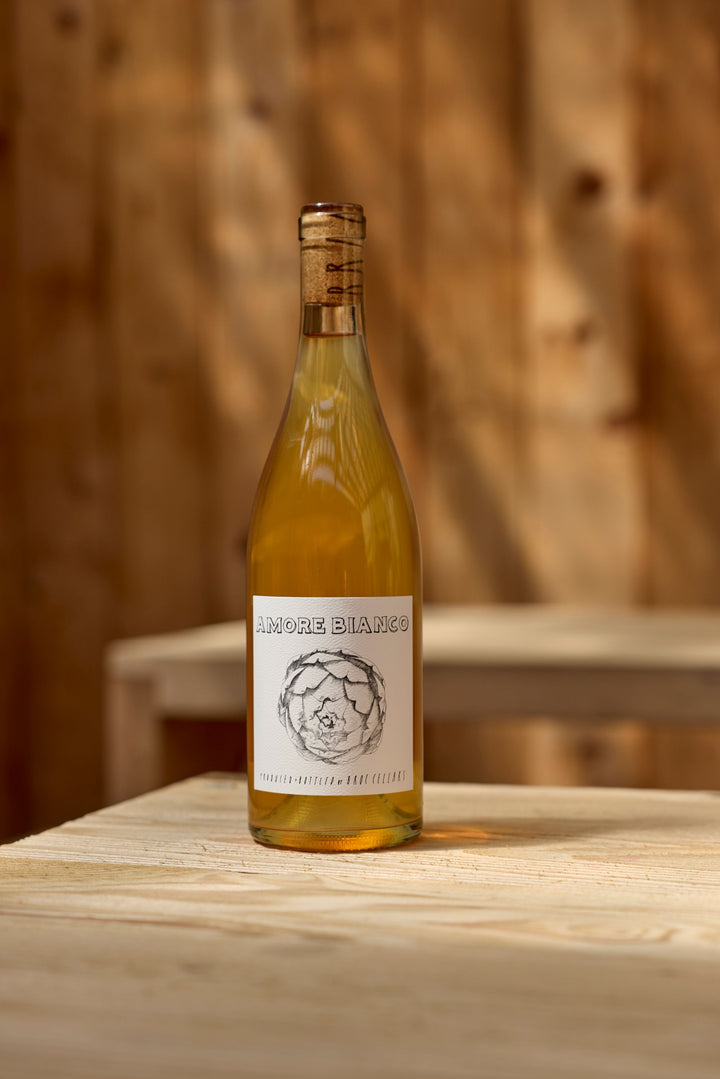 A bottle of 2025 Amore Bianco by Broc Cellars sits on a light wooden table before a wooden wall, its label featuring simple floral artwork and text, all illuminated by natural sunlight.