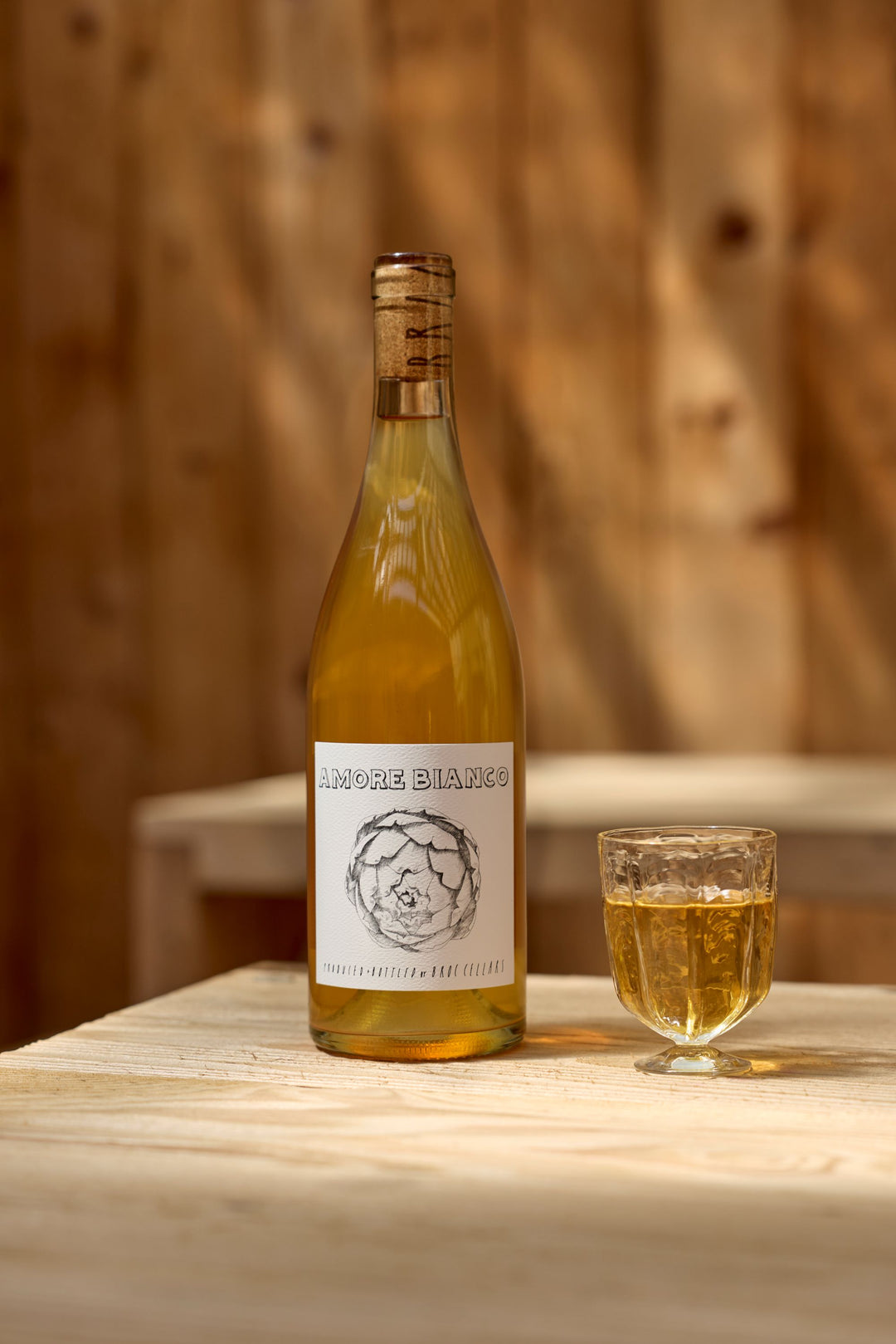 A bottle of 2025 Amore Bianco by Broc Cellars sits beside a glass of pale yellow wine on a wooden table, with a Tech Sheet visible in the blurred wood-toned background.