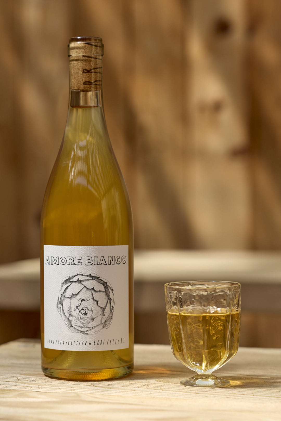 A bottle of Broc Cellars 2025 Amore Bianco skin-contact Tocai Friulano with an artichoke illustration on the label, sitting next to a filled glass.
