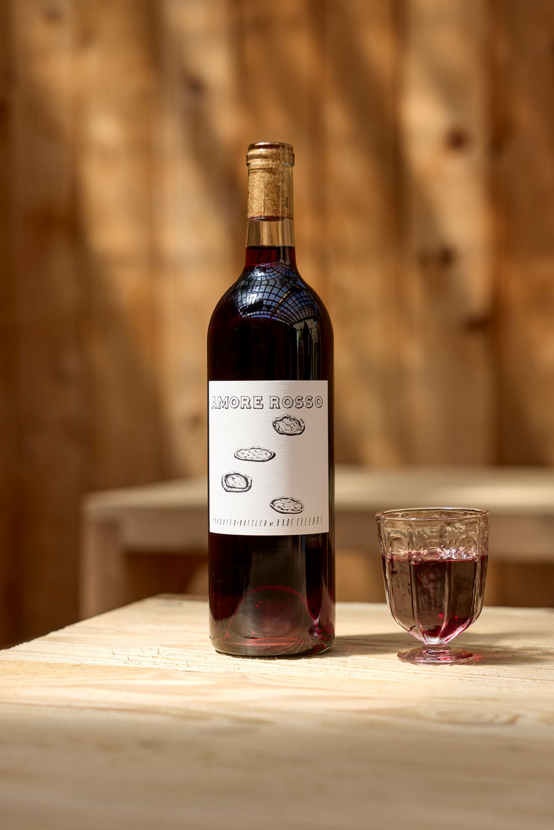A bottle of 2024 Amore Rosso by Broc Cellars sits on a light wooden table beside a glass of red wine, with soft, out-of-focus warm wood paneling in the background. Tech Sheet coming soon.
