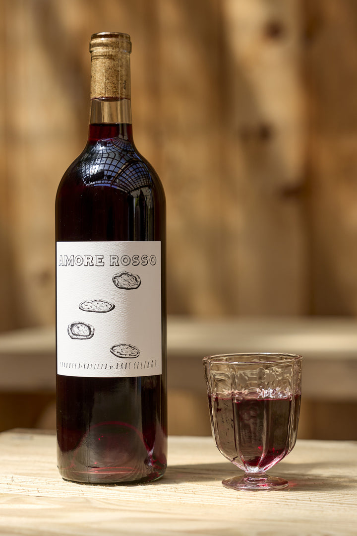 A bottle of 2023 Amore Rosso, a Montepulciano Sangiovese, with a label featuring three circular sketches, standing next to a glass filled with the same red wine.