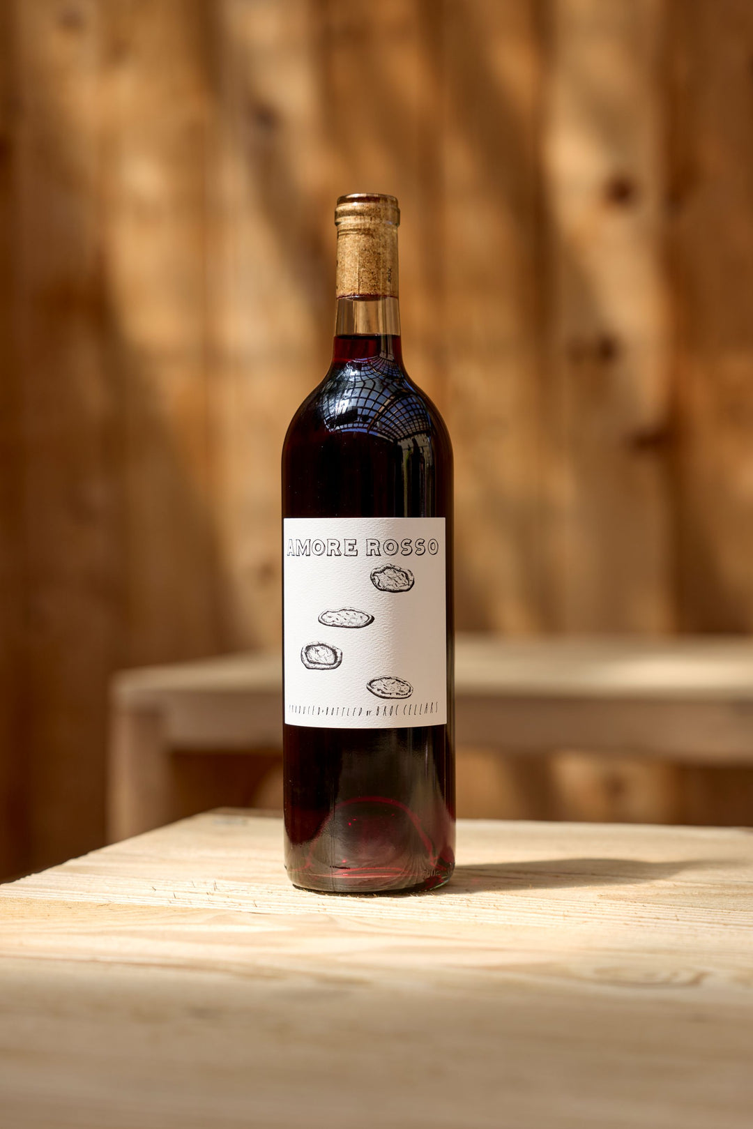 A bottle of 2024 Amore Rosso by Broc Cellars, a classic red wine blend, features simple sketch art on the label as it rests on a light wooden table with a softly blurred wood background.