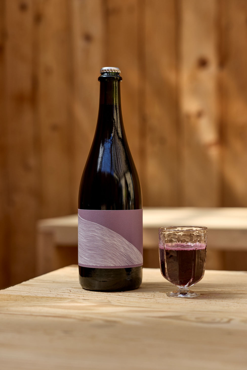 A bottle of Broc Cellars 2024 Aqua Vino sparkling rosé with a purple label sits on a light wooden table next to a glass of pinkish-red wine, set against softly lit wooden planks in the background.