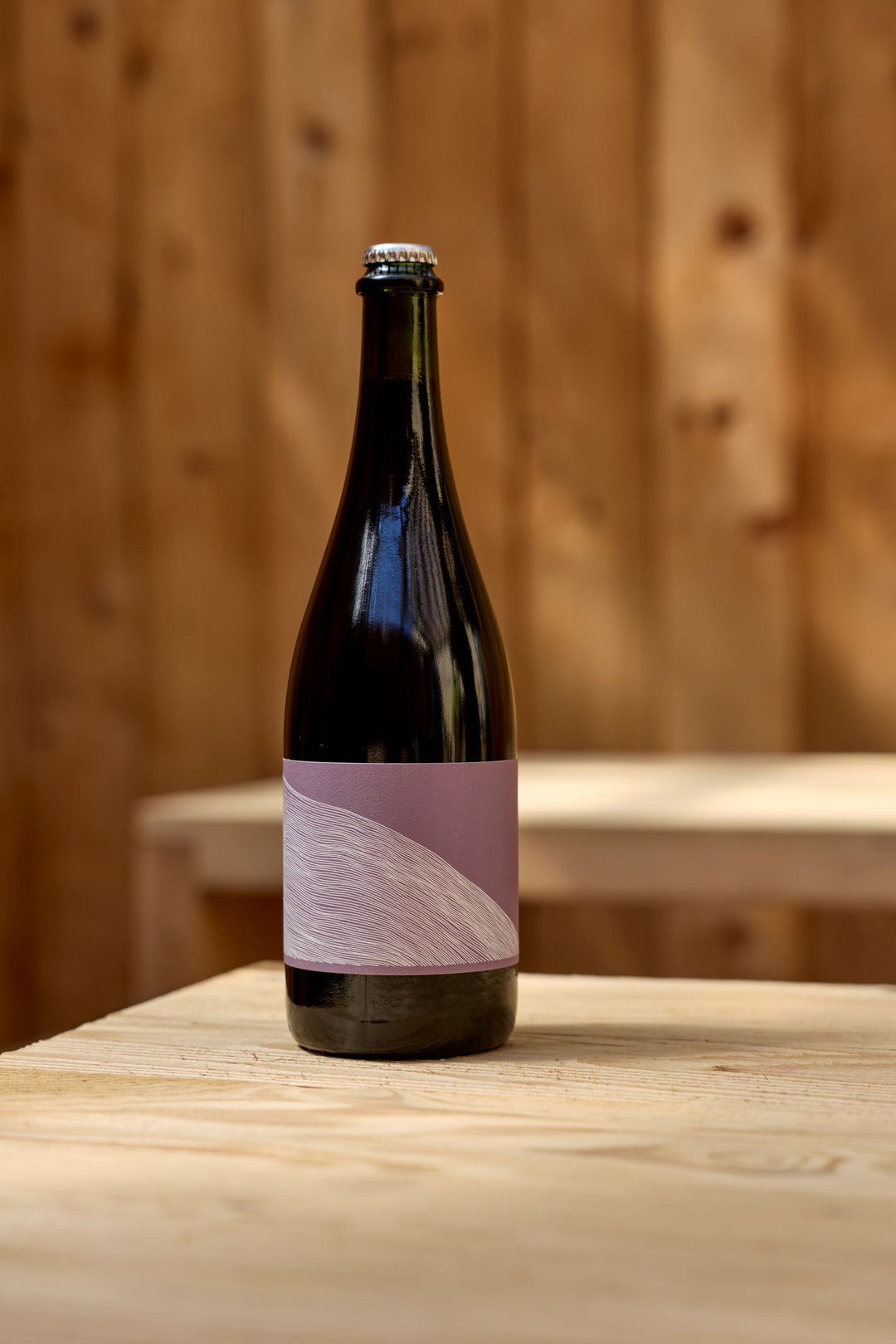 A dark glass bottle of Broc Cellars' 2024 Aqua Vino sparkling rosé with a blank, mauve label rests on a light wooden table, set against a blurred wooden wall backdrop.