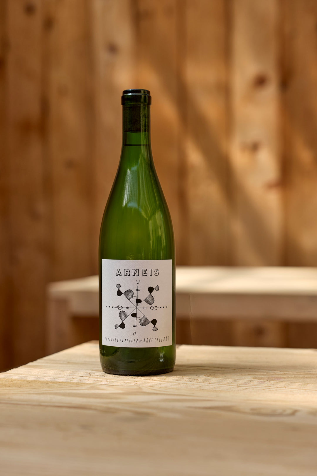 A green wine bottle labeled 2024 Arneis from Broc Cellars sits on a light wood table, with a simple black and white design and a blurred wooden background.