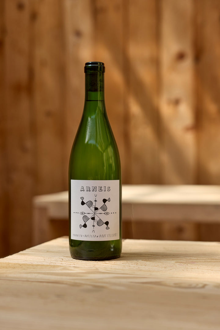 A green wine bottle labeled 2024 Arneis from Broc Cellars sits on a light wood table, with a simple black and white design and a blurred wooden background.