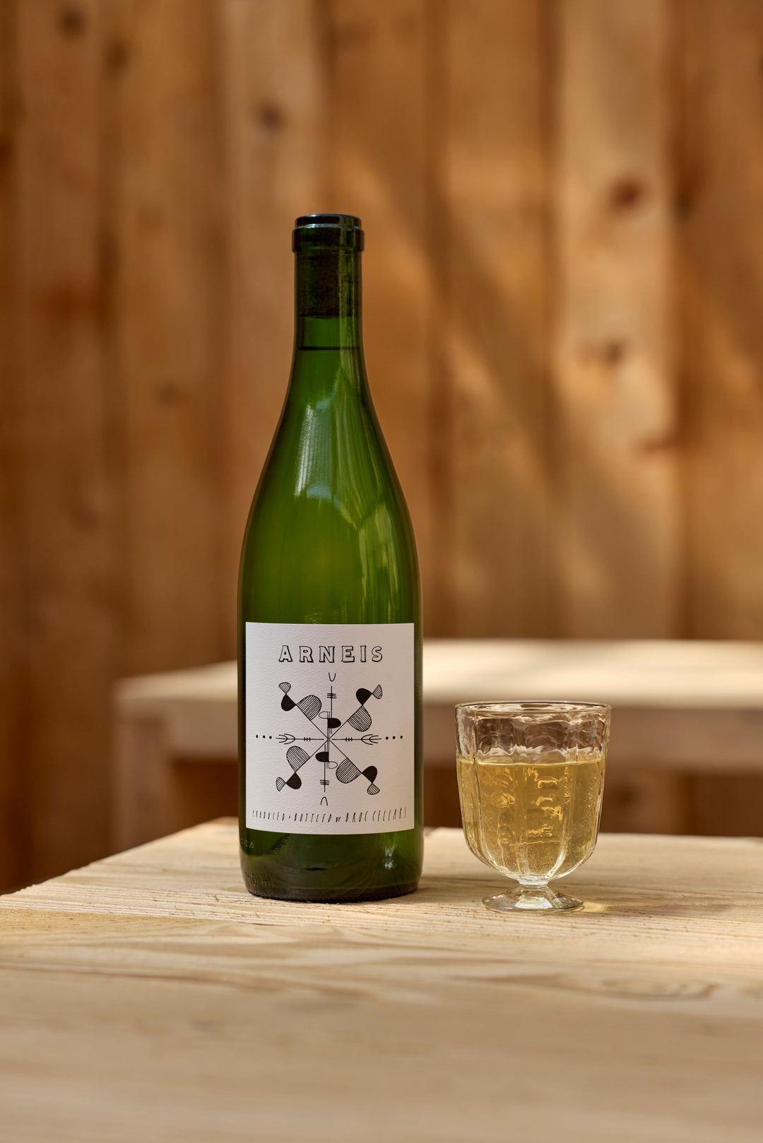 A green bottle of Broc Cellars 2024 Arneis stands beside a glass of white wine on a light wooden table, set against a softly blurred wooden background.
