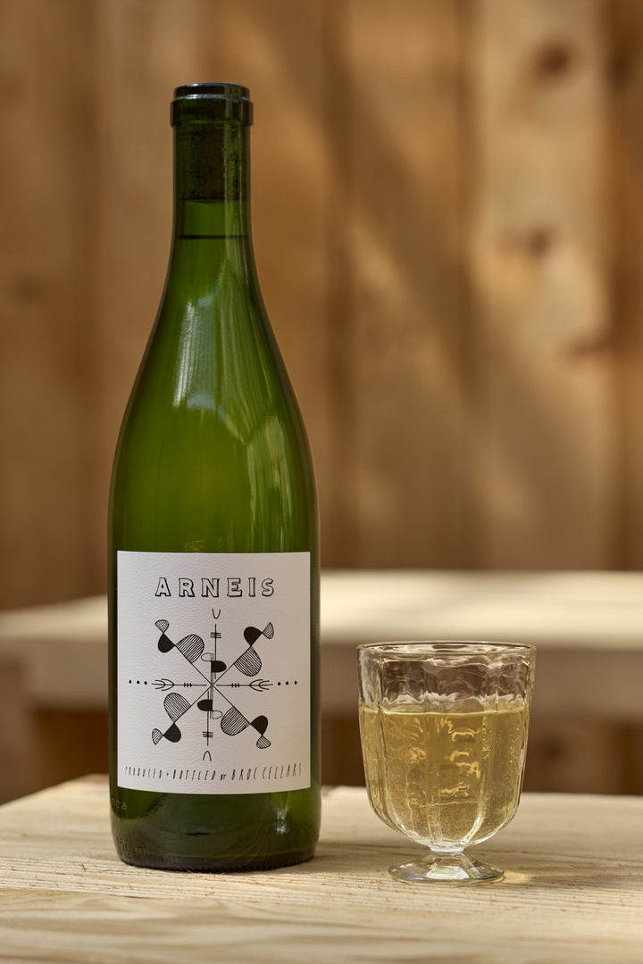 A green glass bottle of Broc Cellars 2024 Arneis with a white label and a small glass of white wine sit on a wooden surface.