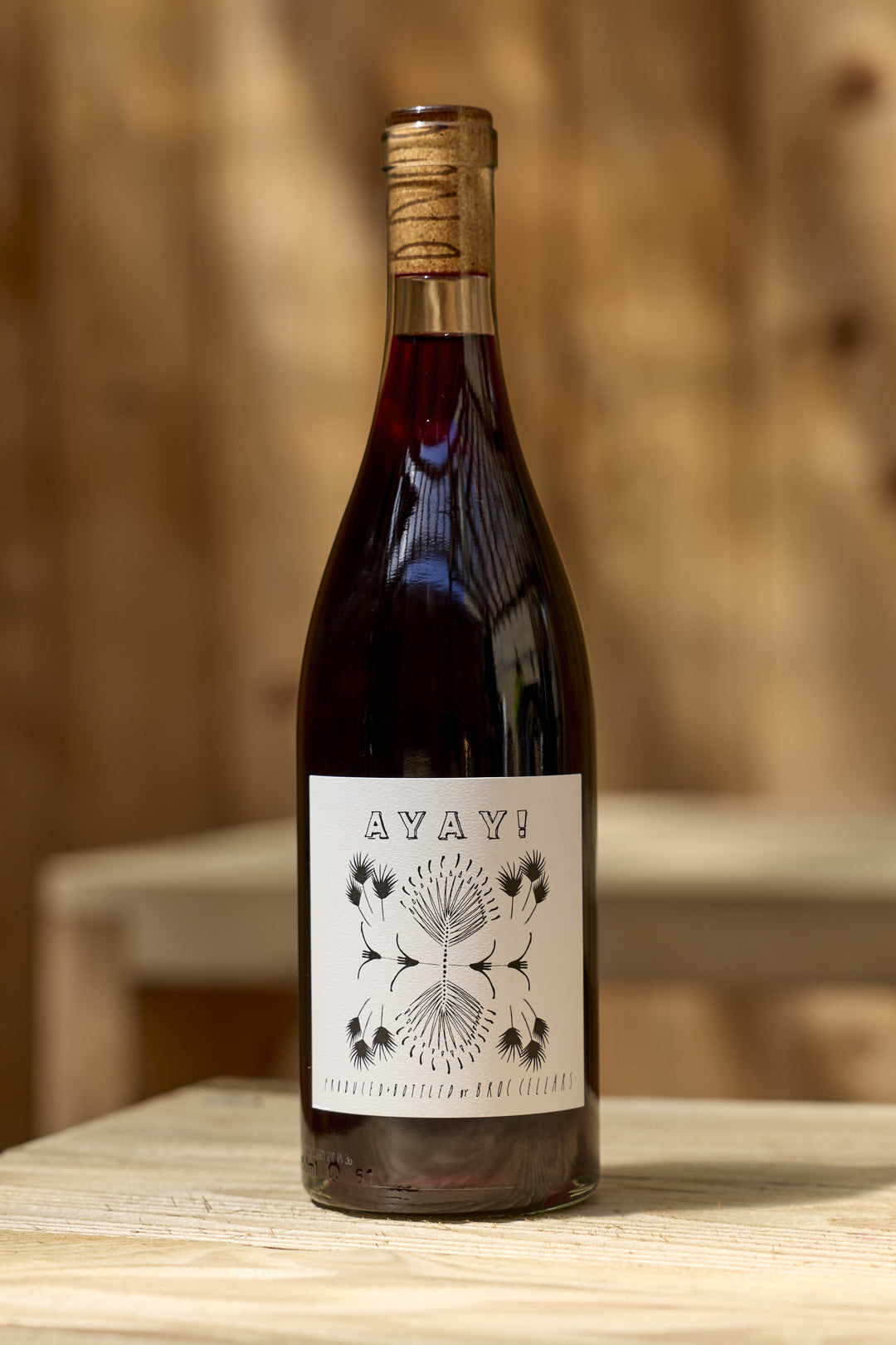 A bottle of Broc Cellars 2025 Ayay! red wine with a white label featuring the text AYAY! above a black-ink line drawing of symmetrical floral shapes.