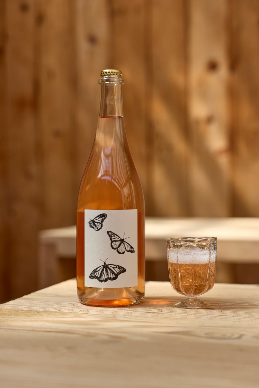 A bottle of Broc Cellars 2025 Sparkling Brachetto, with black and white butterfly illustrations on the label, sits next to a glass of rosé wine on a light wooden table. Wooden paneling is in the background. Coming soon.