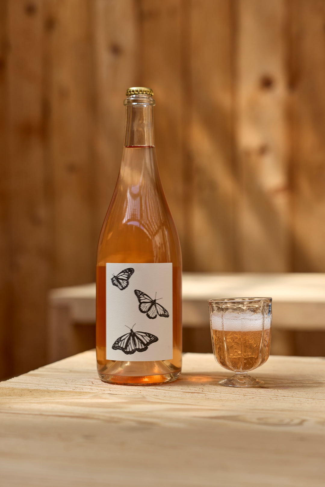 A bottle of Broc Cellars 2025 Sparkling Brachetto, with black and white butterfly illustrations on the label, sits next to a glass of rosé wine on a light wooden table. Wooden paneling is in the background. Coming soon.