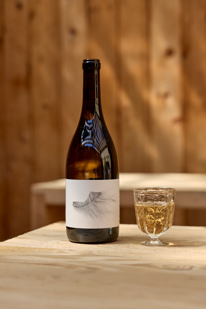 A bottle of Broc Cellars 2023 Michael Mara Chardonnay with a white label featuring a minimalist black line drawing stands next to a glass of white wine on a light wooden table, set against a blurred wood-paneled background.