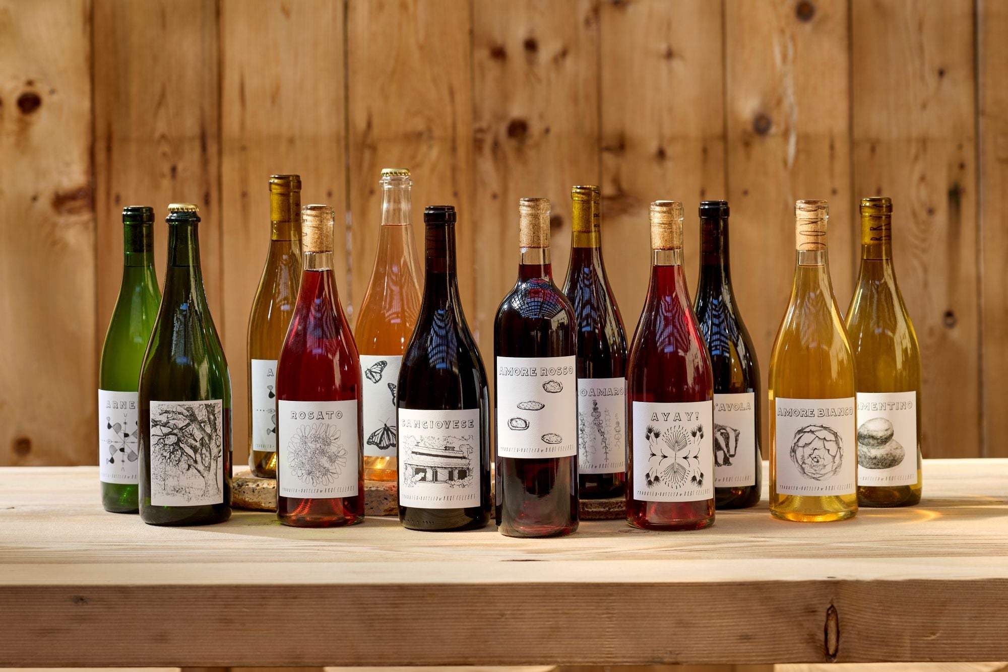 A selection of Fox Hill Collection wines from Broc Cellars—Italian red, rosé, and white wines—feature illustrated labels and cork tops, arranged on a wooden table with a plank wall backdrop.