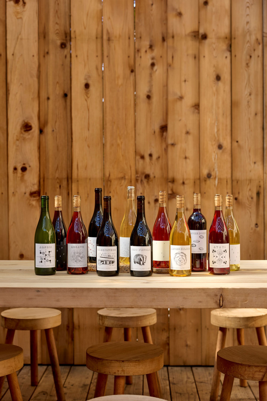 The Broc Collection by Broc Cellars, a 12-bottle wine pack with unique illustrated labels, sits on a wooden table with four stools against a backdrop of vertical wooden planks.