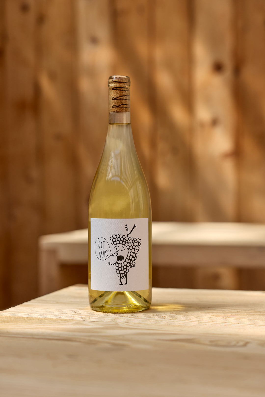 A 2025 Got Grapes White from Broc Cellars with a cork top sits on a wooden table. The label shows a cartoon grape holding a "Got Grapes?" sign, warmly lit against a wooden wall.