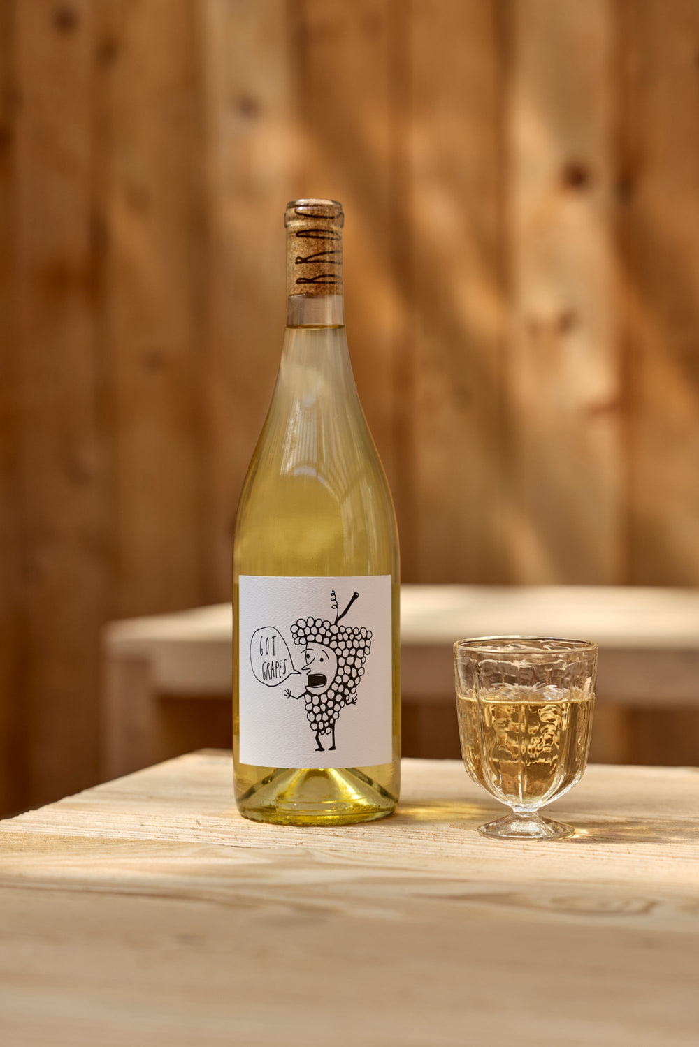 A bottle of Broc Cellars 2025 Got Grapes White, sealed with a cork and sporting a label with a cartoon grape, stands beside a filled glass on a light wooden table, set against a wooden wall backdrop.