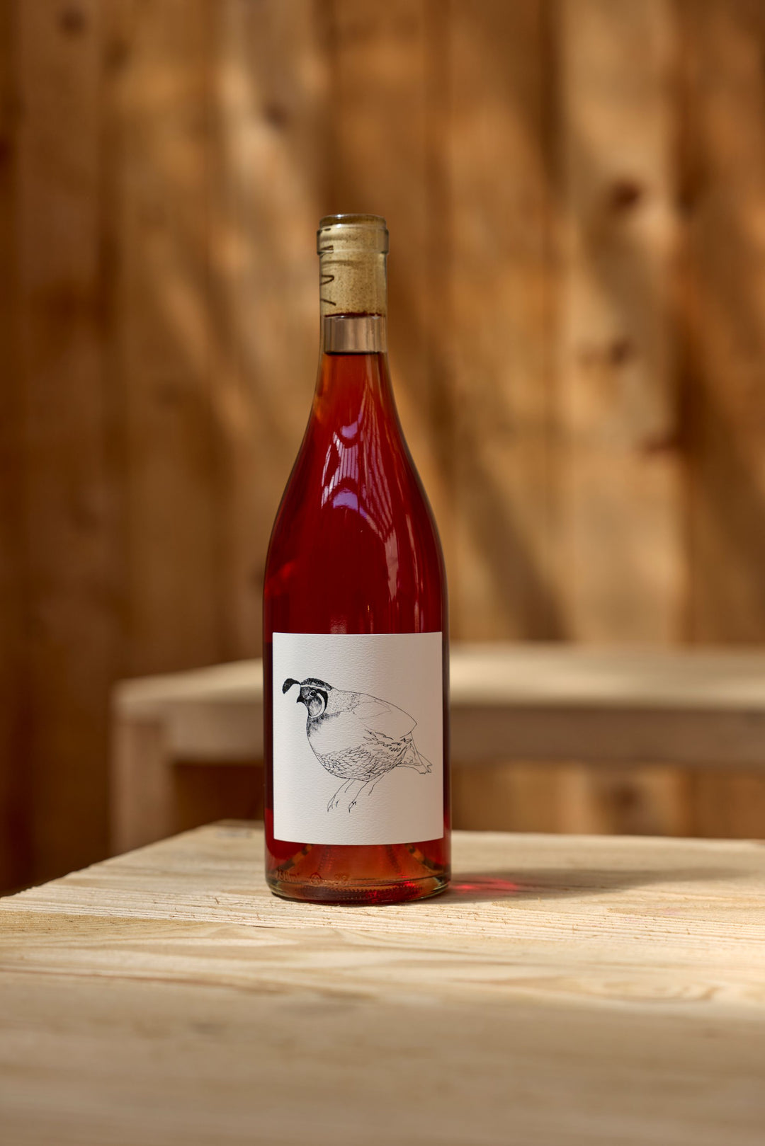 A bottle of Broc Cellars 2023 Grenache Gris rosé with a natural cork rests on a light wooden table. The label shows a minimalist black-and-white quail illustration, with blurred Mendocino County wooden planks in the background.