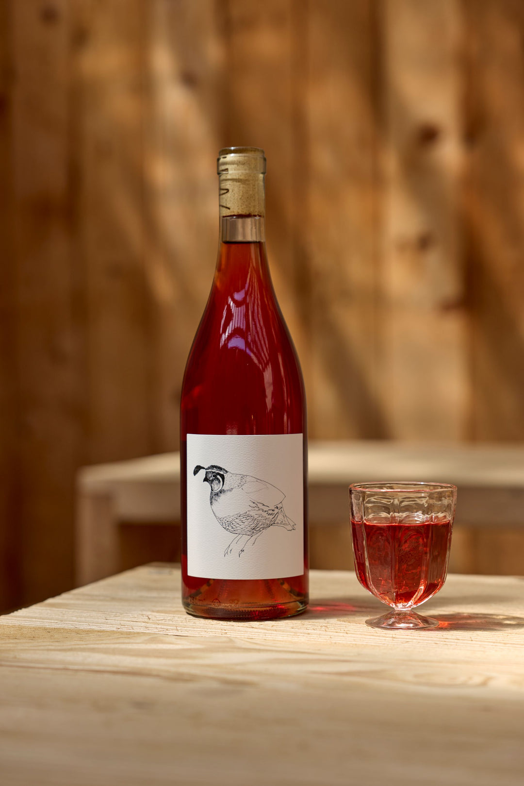 A bottle of Broc Cellars 2023 Grenache Gris, featuring a quail label, stands beside a filled glass on a light wooden table against a wooden wall—crafted in Mendocino County using carbonic maceration.