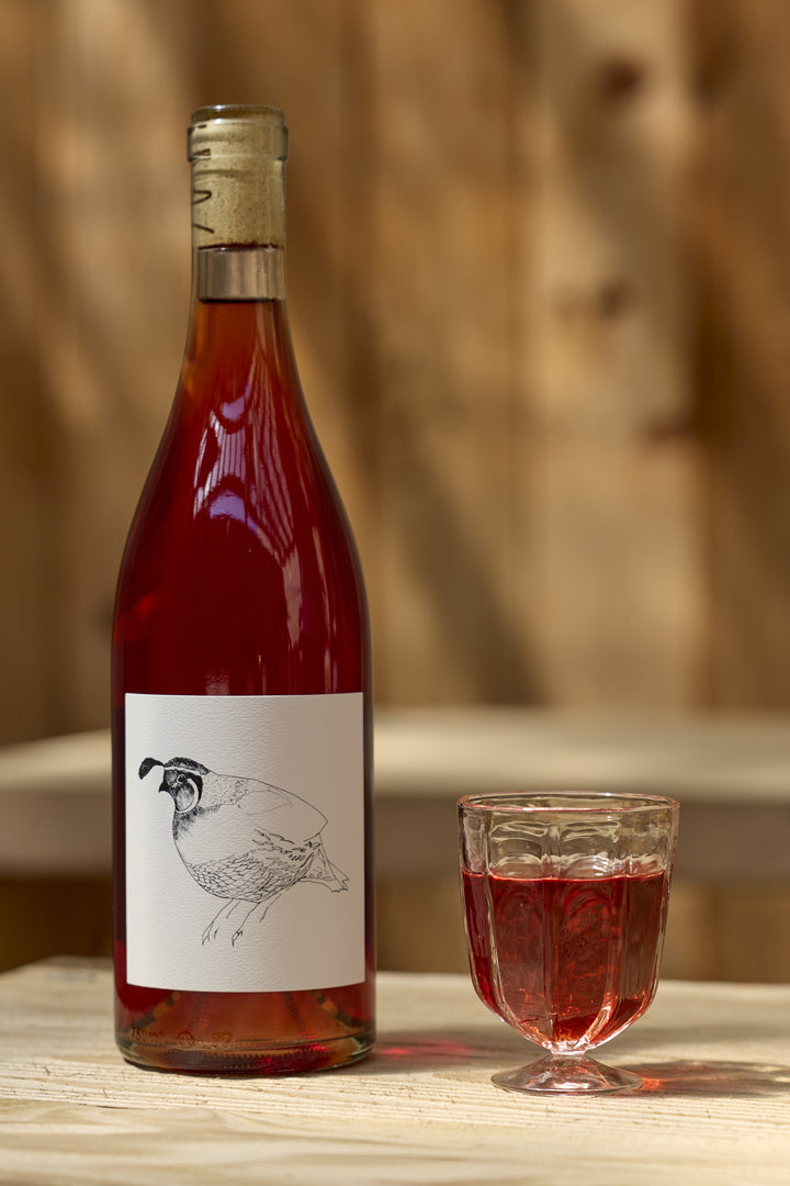 A bottle of Broc Cellars 2023 Grenache Gris rosé featuring a line drawing of a quail on the label, standing next to a small glass of the same wine.