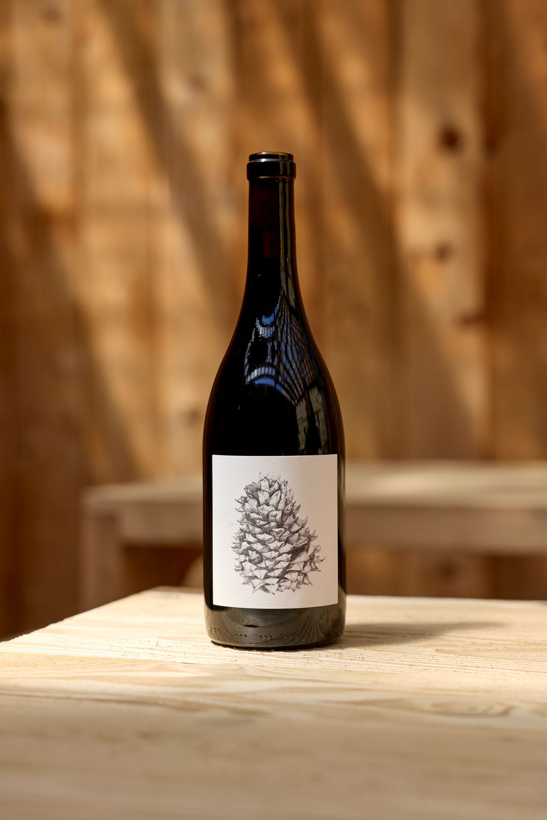 A dark glass bottle of Broc Cellars’ 2024 Lagrein Red, with a white label displaying a detailed black pine cone illustration, sits on a light wooden table against a wooden wall—an ideal image for any tech sheet.