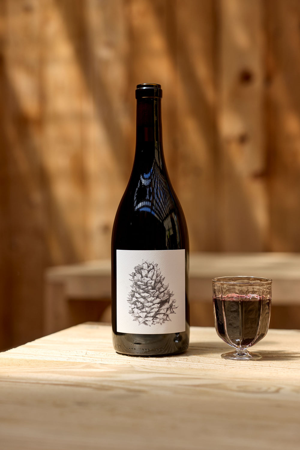 A bottle of Broc Cellars 2024 Lagrein Red with a pine cone label stands beside a glass of red wine on a light wood table, wooden walls behind—an inviting setting to consult the Tech Sheet.