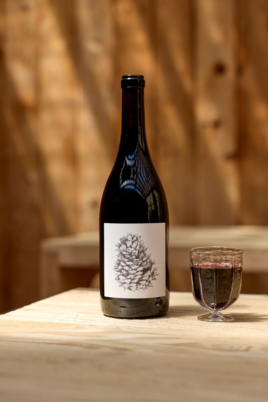 A bottle of Broc Cellars 2024 Lagrein Red with a pine cone label stands beside a glass of red wine on a light wood table, wooden walls behind—an inviting setting to consult the Tech Sheet.