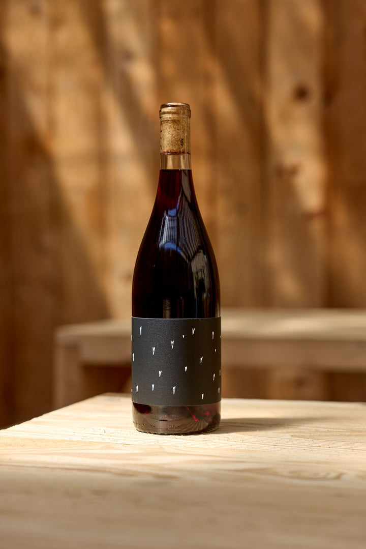 A bottle of Broc Cellars 2023 Love Red, made from California Carignan and sealed with a cork, with a black label featuring small white shapes, sits on a light wooden table against a wooden wall lit by sunlight.