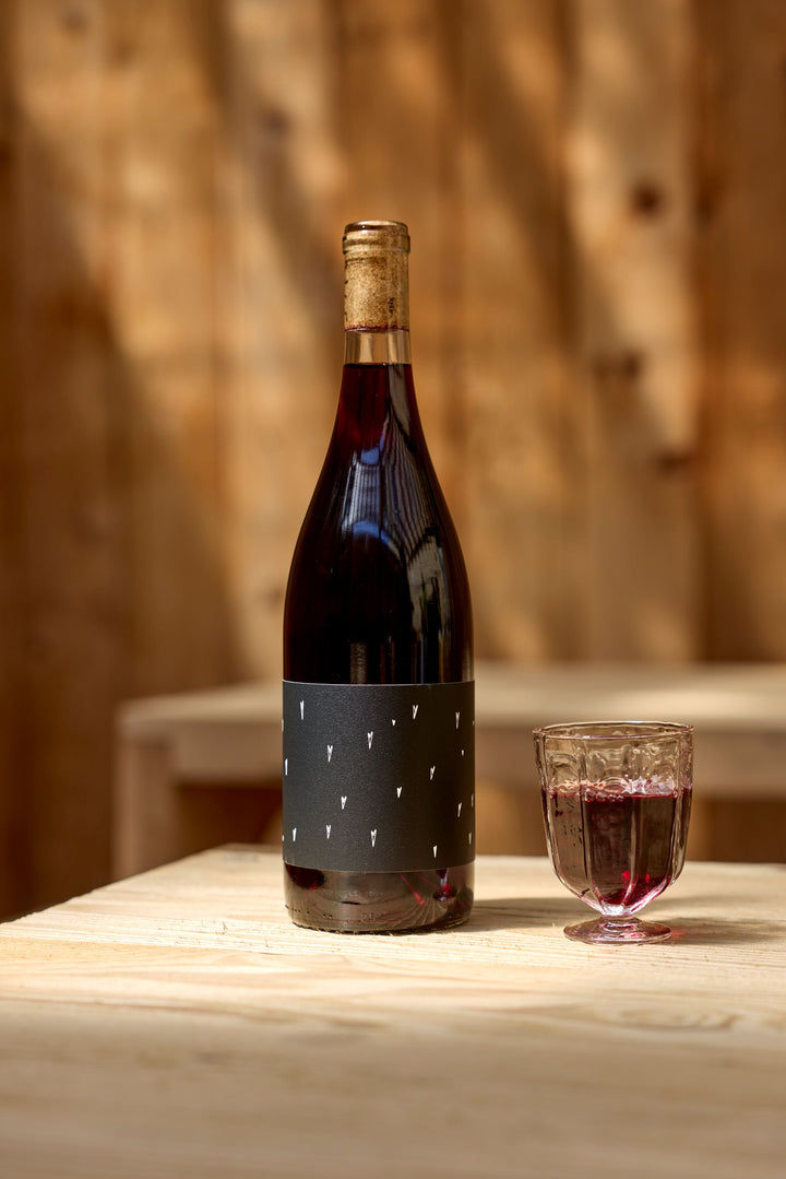 A bottle of Broc Cellars 2023 Love Red, featuring a black label with small white markings, stands on a light wooden table beside a filled glass; a wooden wall is in the background.
