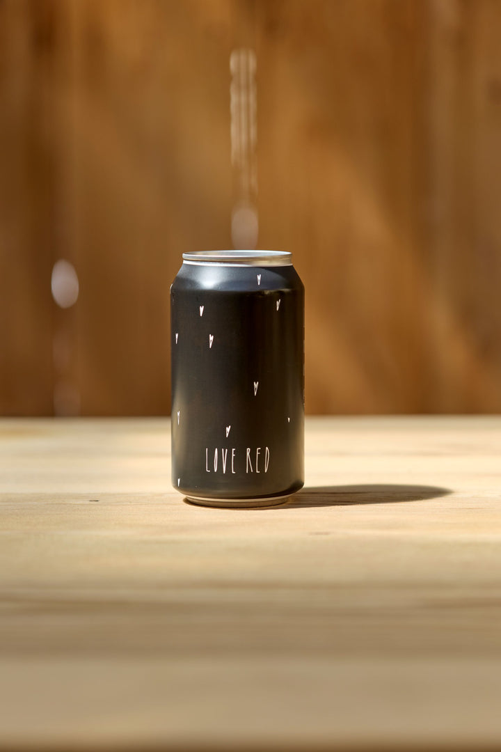 A Love Red Can Pack by Broc Cellars, featuring a black can with small white hearts and LOVE RED text, sits on a light wooden surface, evoking California red wine vibes against a blurred brown background.