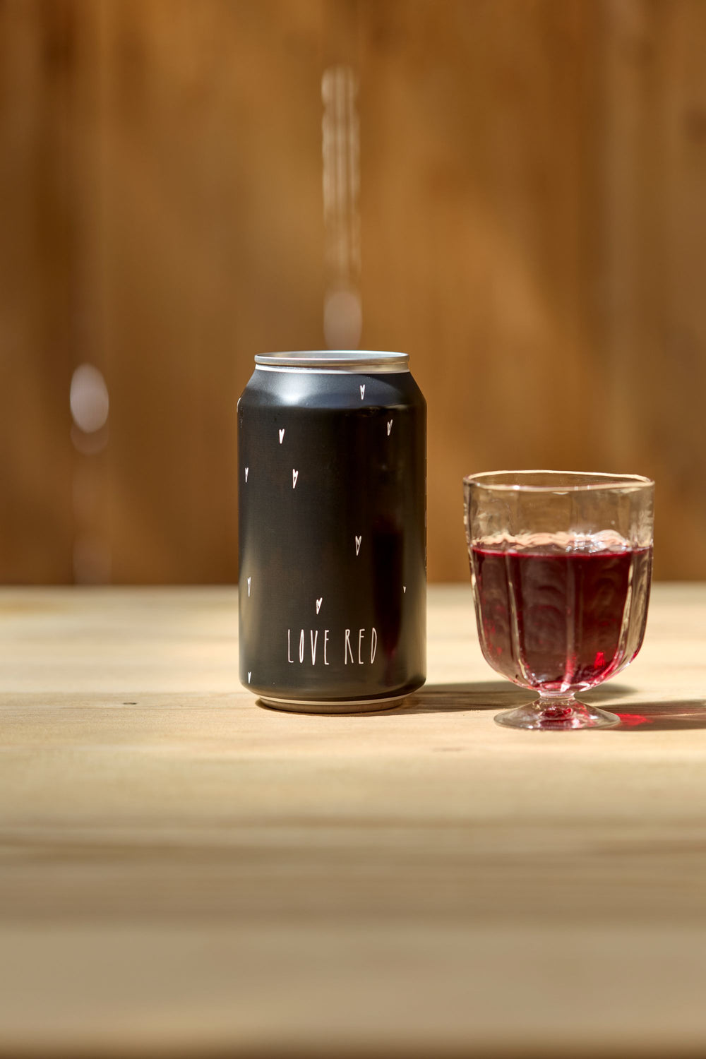 A Love Red Can Pack by Broc Cellars, featuring small heart illustrations, stands next to a glass of red wine on a light wooden surface, set against a blurred brown background.