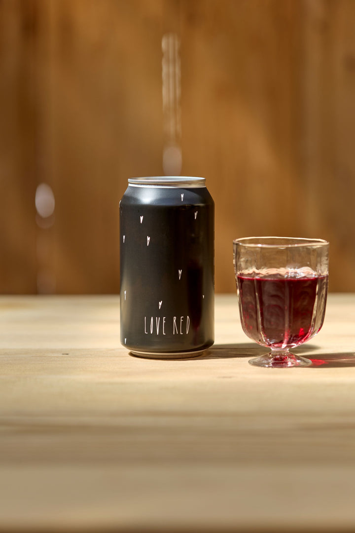 A Love Red Can Pack by Broc Cellars, featuring small heart illustrations, stands next to a glass of red wine on a light wooden surface, set against a blurred brown background.