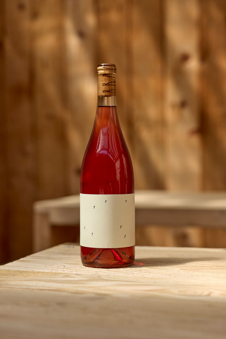 A bottle of 2025 Love Rosé by Broc Cellars, featuring a blank cream label and cork, sits on a light wooden table beside its tech sheet, with a wooden wall and bench in the background.