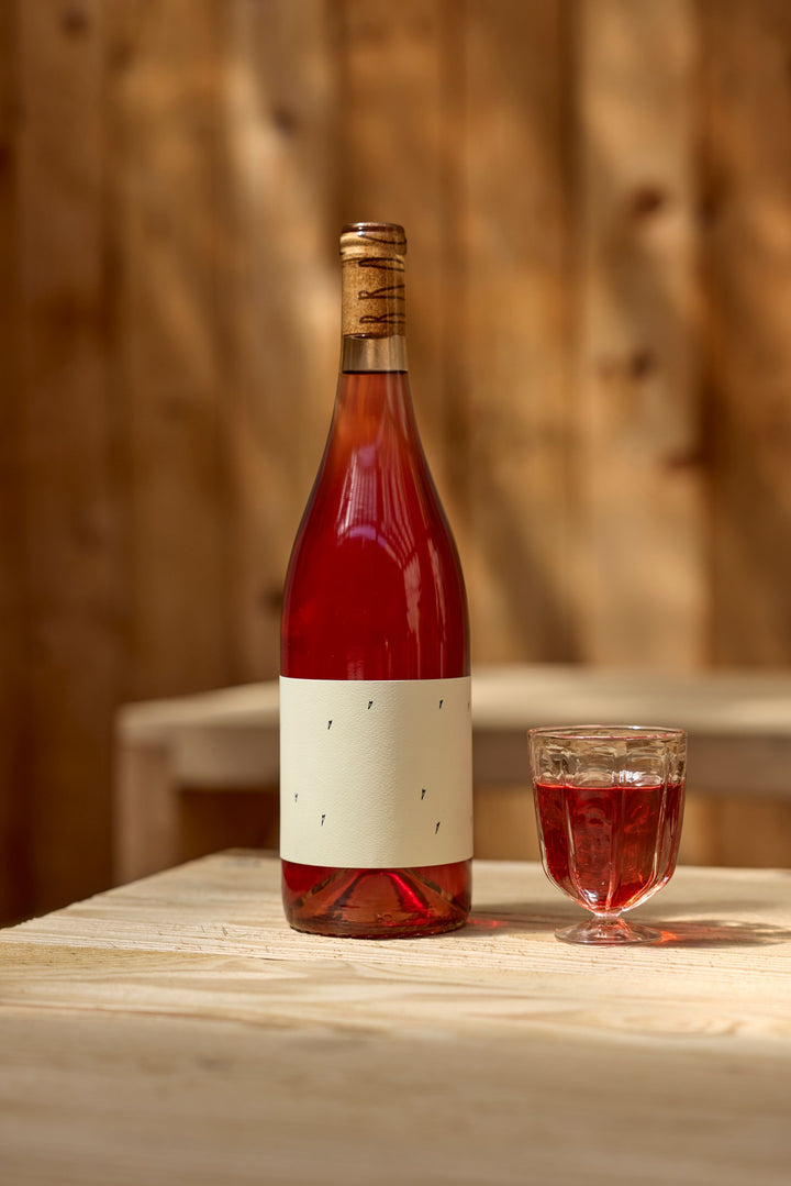 A bottle of Broc Cellars 2025 Love Rosé with a blank cream label stands beside a glass half-filled with rosé on a light wooden table, set against a blurred wood background—ideal for checking the Tech Sheet details.