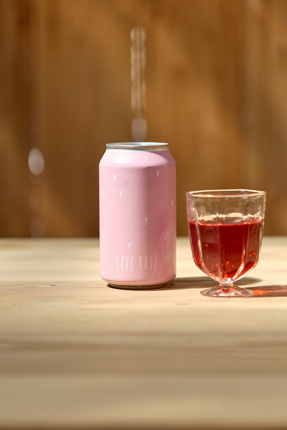 A pink Broc Cellars Love Rosé Can sits next to a glass of pinkish-red Valdiguié rosé, crafted from foot-stomped grapes and natural fermentation, set on a light wooden surface with a blurred brown background.