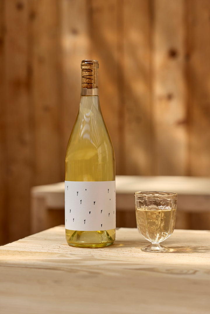 A bottle of Broc Cellars 2025 Love White, crafted from organically farmed grapes, sits on a light wooden table beside a textured glass of white wine, with a softly lit wooden background.