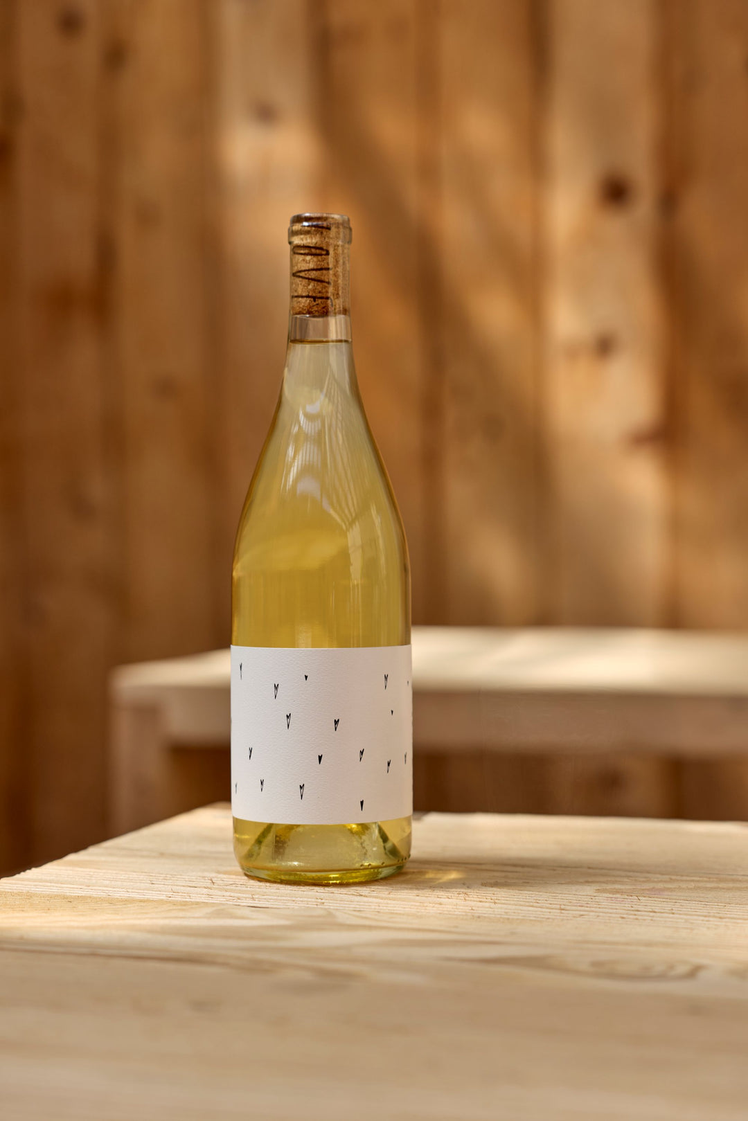 A clear glass bottle of Broc Cellars 2025 Love White, an organic California Vermentino with a cork and minimalist white label, sits on a light wooden table, subtly hinting at its spontaneous fermentation; a blurred wooden wall is in the background.