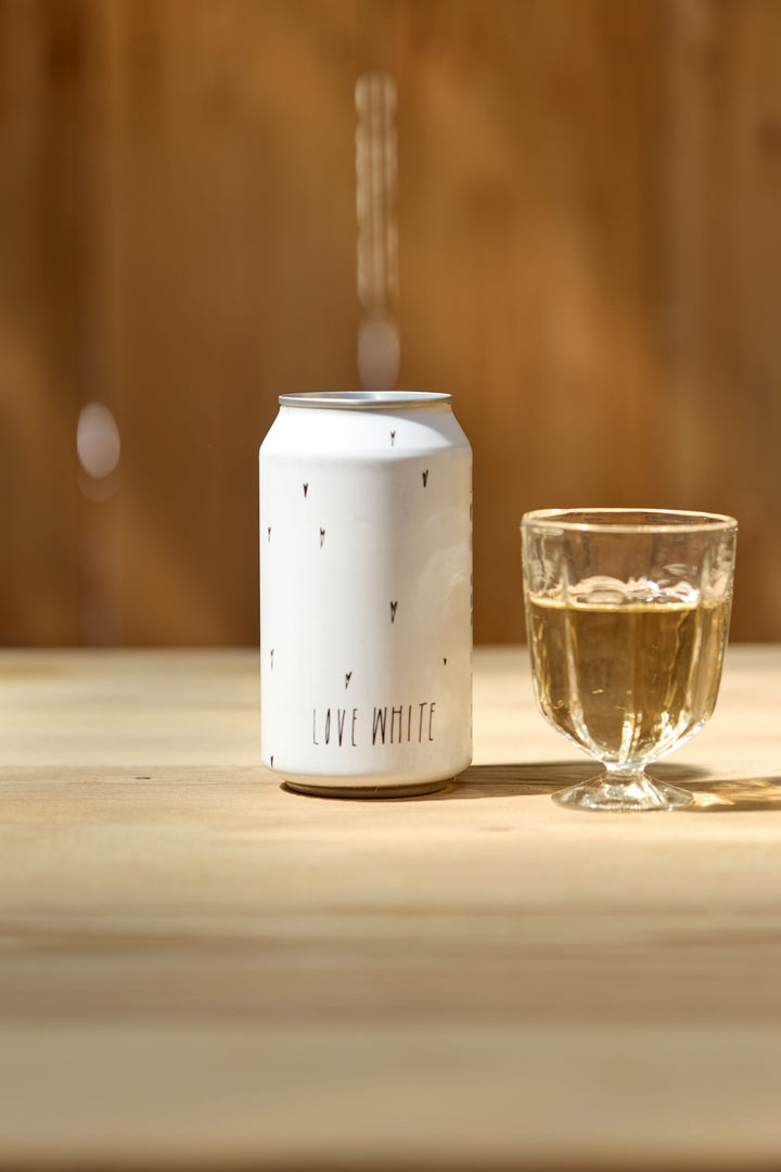 A Broc Cellars Love White Can Pack with small black symbols and "LOVE WHITE" text stands next to a clear glass of light-colored wine, made from organically farmed grapes, atop a wooden surface against a blurred brown background.