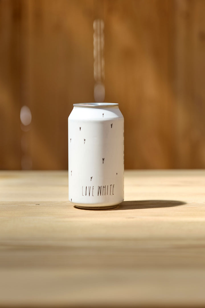 A Broc Cellars Love White Can Pack, decorated with small black triangles and the text LOVE WHITE, rests on a light wooden surface, capturing the refreshing spirit of California white wine made from organically farmed grapes.