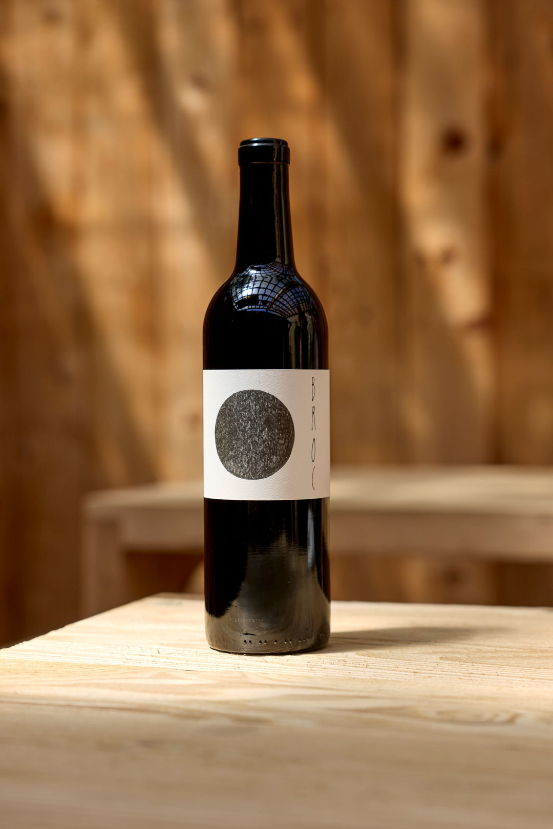 A dark glass bottle of 2023 Broc Massa Cabernet Sauvignon by Broc Cellars, featuring a white label with a large black circle and "BROC" text, rests on a light wooden table against a softly blurred wood background—an elegant organic wine.