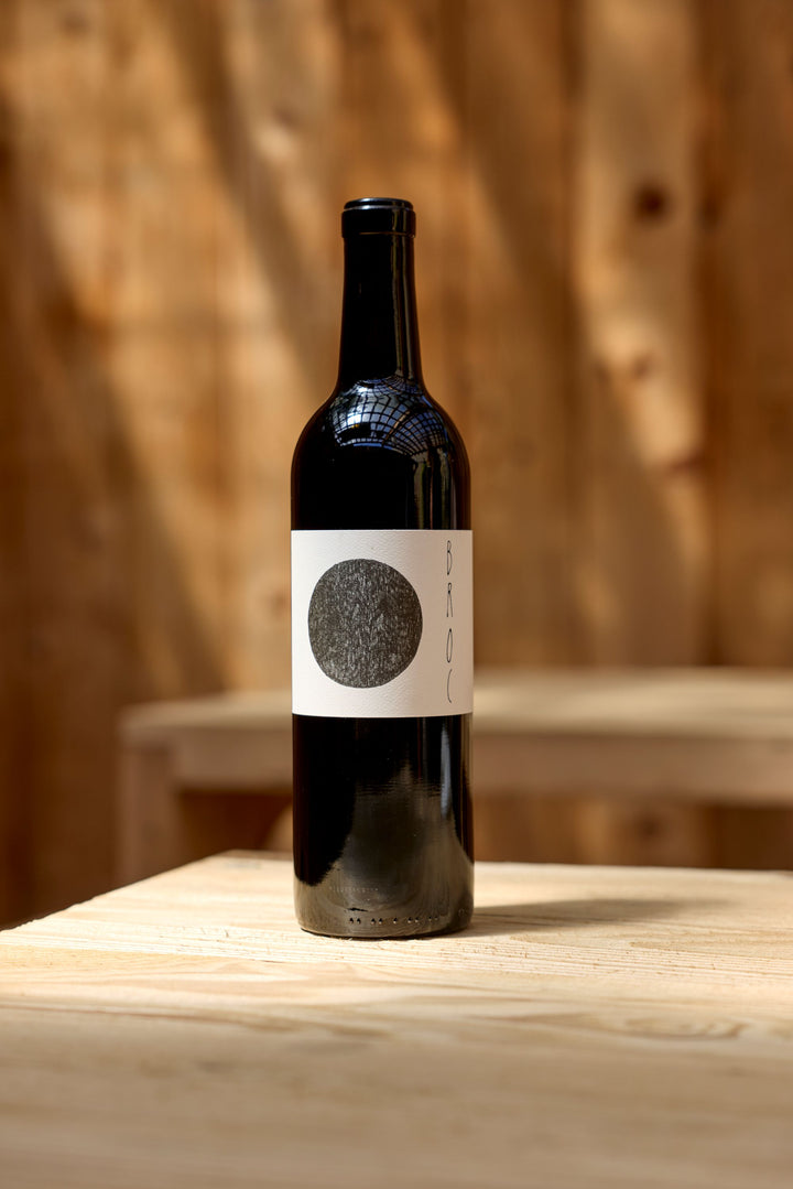 A dark glass bottle of 2023 Broc Massa Cabernet Sauvignon by Broc Cellars, featuring a white label with a large black circle and "BROC" text, rests on a light wooden table against a softly blurred wood background—an elegant organic wine.