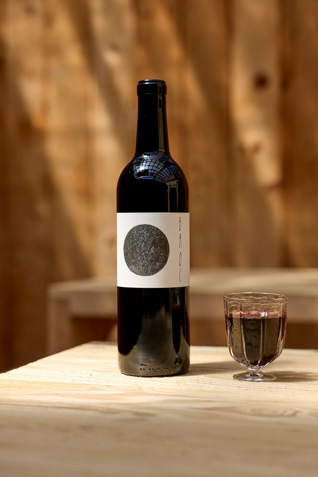 A bottle of 2023 Broc Massa Cabernet Sauvignon from Broc Cellars, with a white label and black circle, sits beside a glass of red wine on a light wooden table against a blurred wooden background.